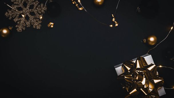 Weihnachtsgeschenk. Weißes Geschenk mit goldener Schleife, goldenen Kugeln und funkelnden Lichtergirlanden in Weihnachtsdekoration auf dunklem Hintergrund zur Grußkarte. Weihnachtskulisse mit Platz für Text - Foto, Bild