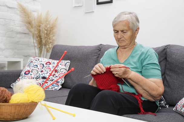 ソファに座り、赤いウールで編むシニア女性。手作りのウールアイテム。クラフト& DIYのコンセプト. - 写真・画像