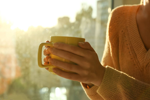 Woman holding cup with tea near window at home, closeup - Foto, afbeelding