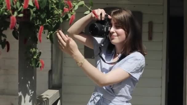 Una joven que ríe se para en la puerta de un café de verano con una cámara y toma fotos de una planta interesante mientras la sostiene con la mano. Vista frontal. Plan medio - Imágenes, Vídeo