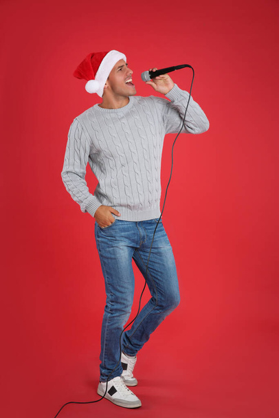 Happy man in Santa Claus hat singing with microphone on red background. Christmas music - Фото, зображення