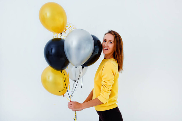 A young woman in a yellow sweater holds a bunch of gold, white and silver balloons against a white wall - Photo, Image