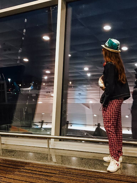 À espera de um voo atrasado no aeroporto à noite. Uma turista indo de férias, de pé no aeroporto e olhando para a janela em aviões. Foto por trás. - Foto, Imagem