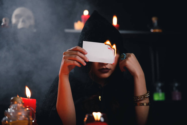 Fortune teller σε μαύρο ρόμπα κρατώντας έγκαυμα χαρτί με τον καπνό ως φόντο. - Φωτογραφία, εικόνα