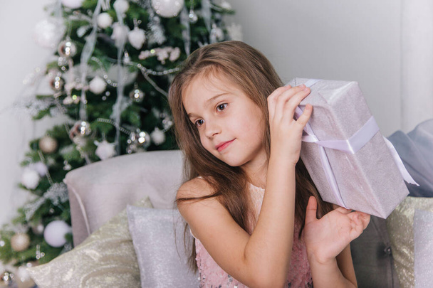 エレガントな光沢のあるドレスに長い髪を持つ幸せな女の子は彼女の手に贈り物を保持しています。クリスマスの背景の上の子供. - 写真・画像