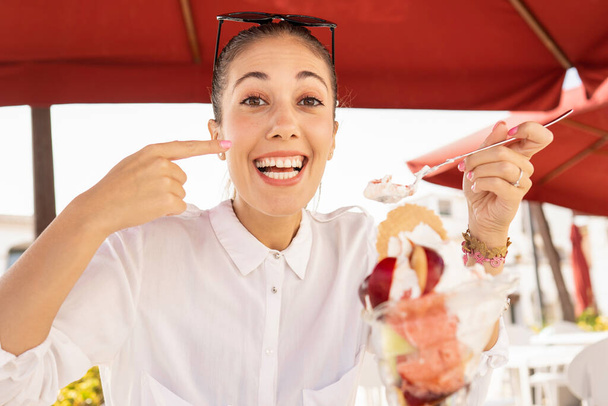Piękny uśmiech szczęśliwy młoda kaukaska kobieta robi pyszny gest palcem podczas jedzenia dużych złożonych lodów z owocami - Nowe pokarmowe złe nawyki ludzi obżarstwo - Zdjęcie, obraz