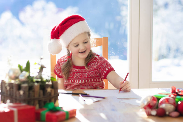 Kind schreibt Brief an Weihnachtsmann an Heiligabend. Kinder schreiben Weihnachtswunschzettel. Kleines Mädchen sitzt im geschmückten Wohnzimmer mit großem Fenster an einem sonnigen Wintertag in den Bergen, draußen schneit es. - Foto, Bild
