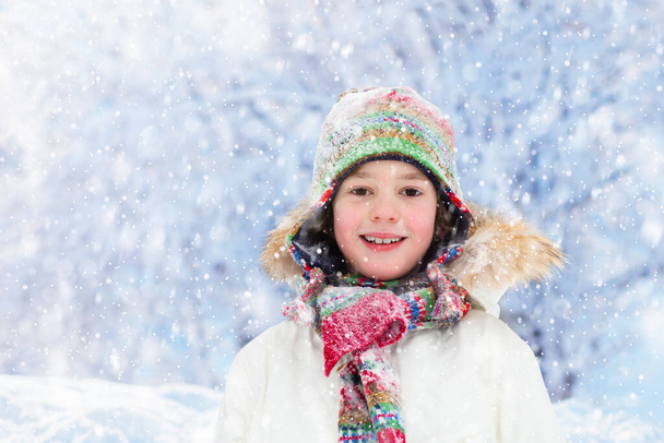 Dítě si v zimě hraje se sněhem. Malý chlapec v pestré bundě a pletený klobouk chytání sněhové vločky v zimním parku na Vánoce. Děti si hrají a skákají v zasněženém lese. Sněhová koule boj pro děti. - Fotografie, Obrázek