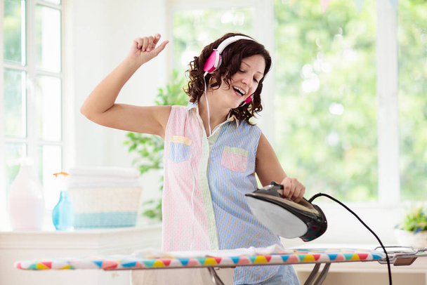 Woman ironing clothes, listening to music and dancing. Young female in headphones folding clothes at iron board. Dance and sing, home chores fun. Housewife cleaning house. - Photo, Image