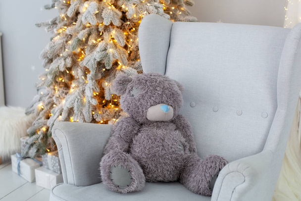 Teddy bear sitting on grey armchair by glowing lights Christmas tree with gifts. Scandinavian decor xmas child room. New Year. Cozy Xmas interior home. Baby room interior. Christmas present. family - Photo, Image