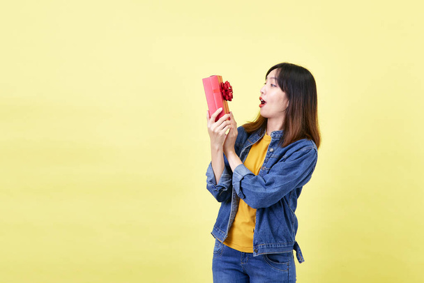 Um feliz animado bela mulher asiática na mão abrindo uma caixa de presente vermelha no estúdio contra um fundo amarelo - Foto, Imagem