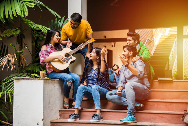 Asian Indian college students playing music with guitar while sitting in campus on stairs or over lawn - Photo, Image