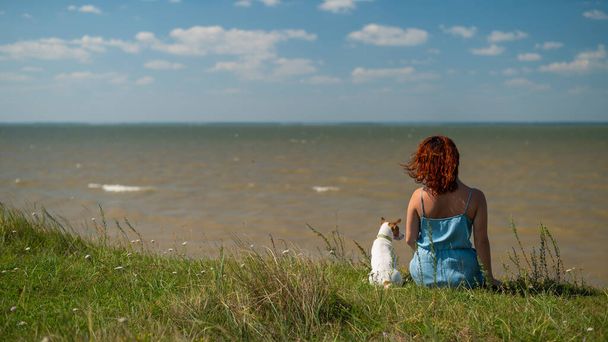 A woman sits on the shore of a pond with a dog. Back view of a girl on vacation in nature with a pet. - Photo, Image