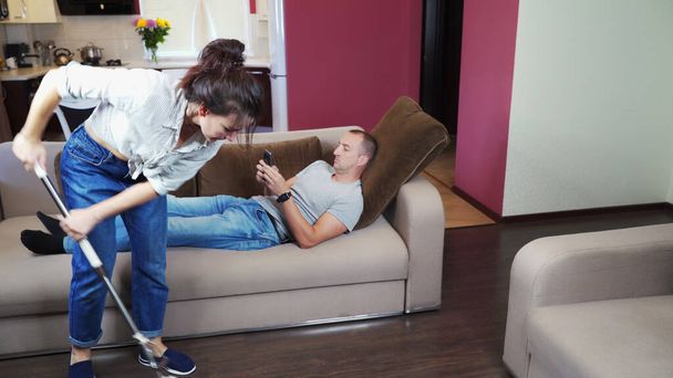 A man indifferently rests on the couch and looks at his smartphone while the girl does the cleaning - Valokuva, kuva