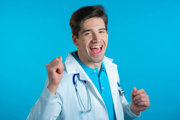Smiling doctor funny dancing, success and luck at work. Young handsome doc man in professional medical white coat is isolated on blue studio background. - Fotó, kép