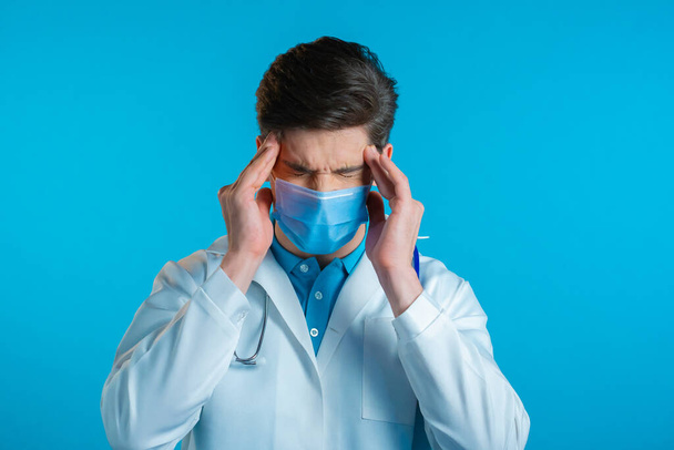 Young doctor man in professional medical white coat and mask having headache, studio portrait. Guy putting hands on head, isolated on blue background. Concept of problems, medicine, illness - Foto, Bild