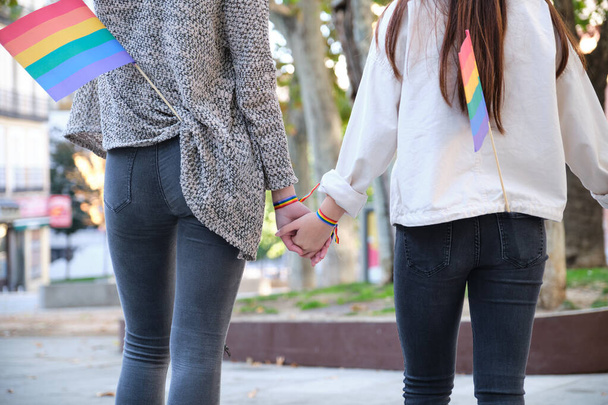 Pareja lesbiana irreconocible cogida de la mano con pulseras LGBT y banderas LGBT en los bolsillos. Celebración del orgullo LGBT. - Foto, Imagen