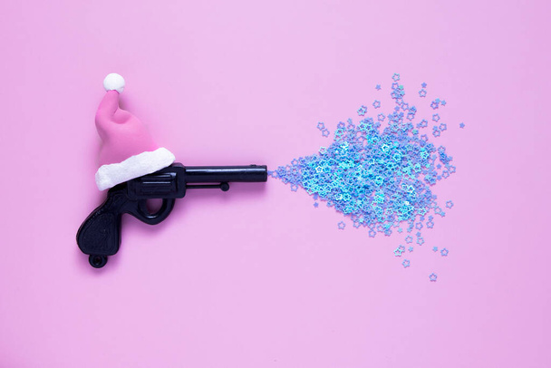 креативная новогодняя концепция: рождественский игрушечный пистолет в шляпе Санты с мини-звездами на розовом фоне - Фото, изображение