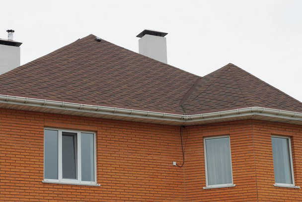 кирпичный дом с белыми окнами и коричневой черепичной крышей с дымоходами против серого неба - Фото, изображение