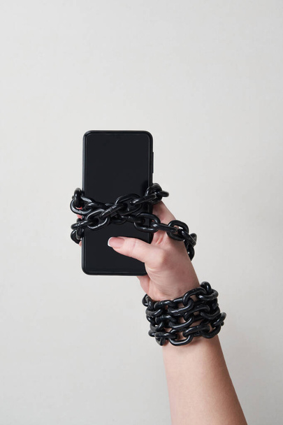 IJzeren ketting die hand en smartphone verbindt in concept van sociale media en internetverslaving op grijze achtergrond - Foto, afbeelding