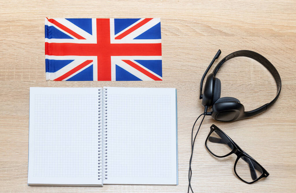 Internet learning of English. Top view of headphones with a microphone for online learning, a notebook for notes and glasses along with the flag of Great Britain. - Foto, Bild