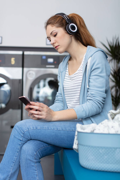 happy woman listens to music on headphones in laundry room - Φωτογραφία, εικόνα