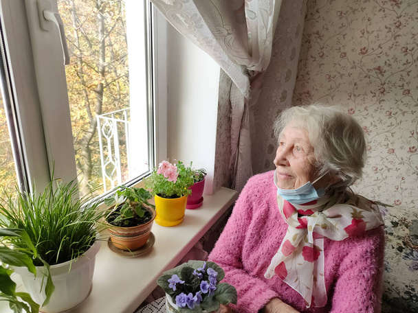 Druga fala. Covid-19, koncepcja zdrowia, bezpieczeństwa i pandemii - starsza samotna kobieta nosząca ochronną maskę medyczną siedząca w pobliżu okna w domu dla ochrony przed wirusem - Zdjęcie, obraz