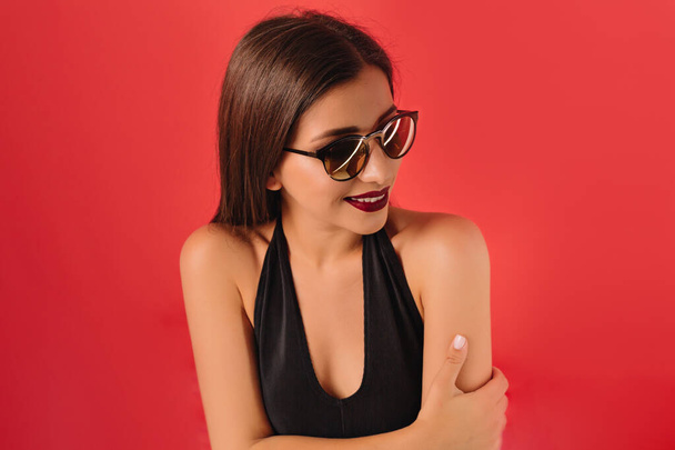 Mujer de moda con el pelo largo con vestido negro y gafas de sol con los labios rojos de pie sobre el fondo rojo. Foto de alta calidad. - Foto, Imagen