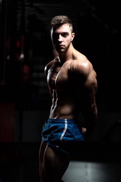Hombre joven de pie fuerte en el gimnasio y músculos flexionantes - Muscular atlético culturista modelo de fitness posando después de los ejercicios - Foto, Imagen
