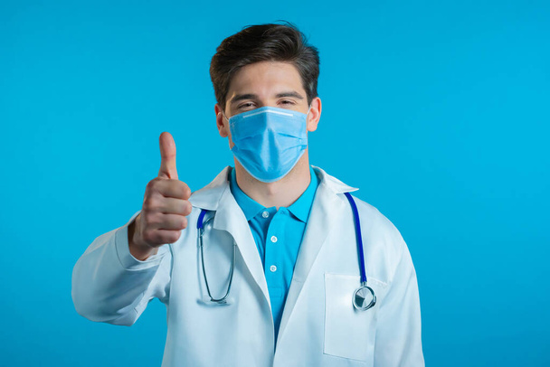 Doctor in mask showing thumb up sign over blue background. Positive young man in medical coat smiles to camera. Winner. Success. Body language. - Фото, изображение