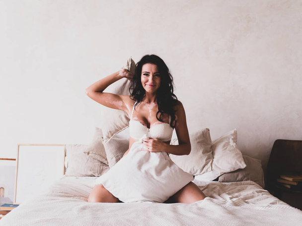 枕付きの白い寝室で白い下着に白のロゴと暗い髪の魅力的な女性の肖像画。不完全な完全性の概念。朝の概念 - 写真・画像