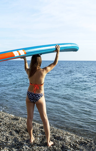 Fiatal, sportos, kaukázusi nő hátulról bikiniben, kezében eveződeszkával a feje fölött a kék tenger partján. Eveződeszka, aktivitás, sup, vízi sportok - Fotó, kép