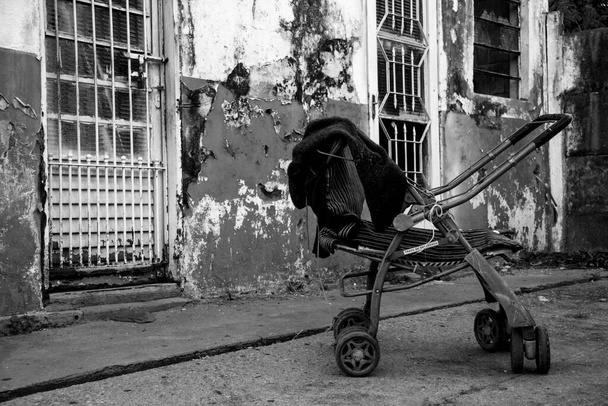 Cochecito abandonado cerca de casa degradada con puertas de hierro y ventana en una zona marginal o pobre de la ciudad (colores blanco y negro)) - Foto, imagen