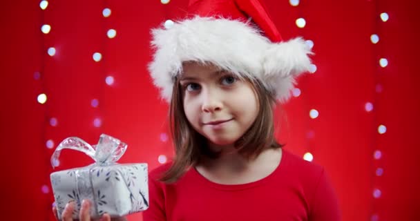 cute girl in a santa hat holds a gift on a red christmas background - Imágenes, Vídeo