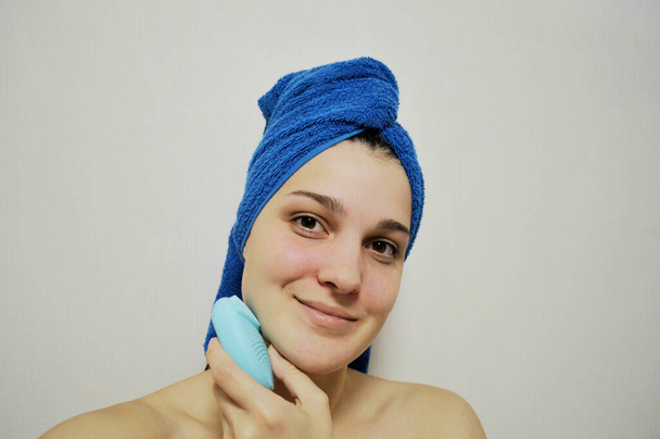 Kafasında mavi havluyla arka planda oturan kadın yüzünü elektrikli fırçayla temizliyor. - Fotoğraf, Görsel