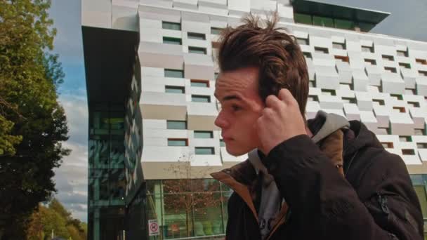 Junger Mann telefoniert mit drahtlosen Kopfhörern im Freien, während er die Straße hinuntergeht. junger Kerl, Teenager trägt moderne drahtlose Kopfhörer. Porträt eines jungen Mannes. - Filmmaterial, Video