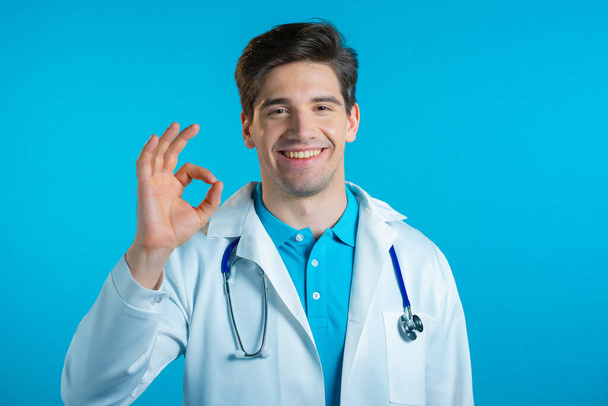 Man in medical white coat showing OK sign over blue background. Positive doctor smiles to camera. Winner. Success. Body language. - Foto, afbeelding