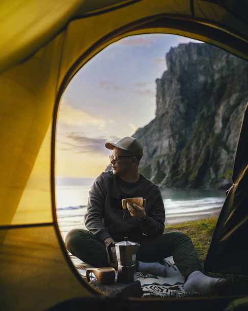 Camping with tent in Lofoten islands, Northern Norway, Kvalvika beach, during sunset. Person sitting in front of a tent holding a hot drink mug. - Фото, зображення