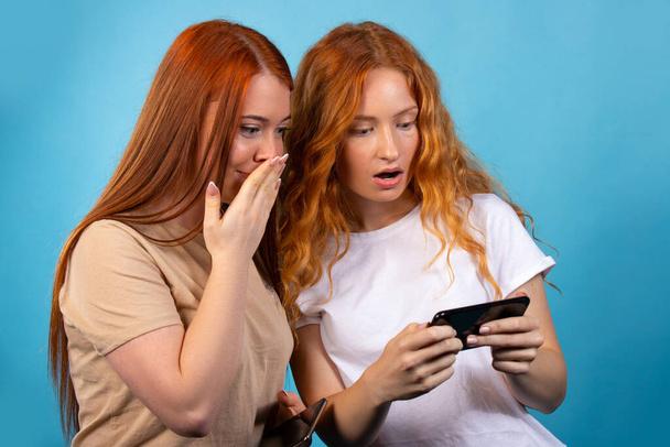 Two red-haired girlfriends in casual clothes. Isolated on blue background. Surprised looking at something on the phone. People lifestyle concept. High quality photo - Foto, Imagen