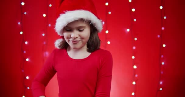 cute girl in a santa hat opens a gift on a red Christmas background - Imágenes, Vídeo