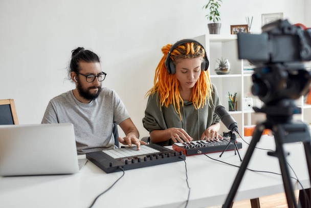 Creative process. Woman with dreadlocks singing and playing. Female and male blogger making music using synthesizer, drum pad machine and laptop, recording video blog or vlog at home - Foto, Bild