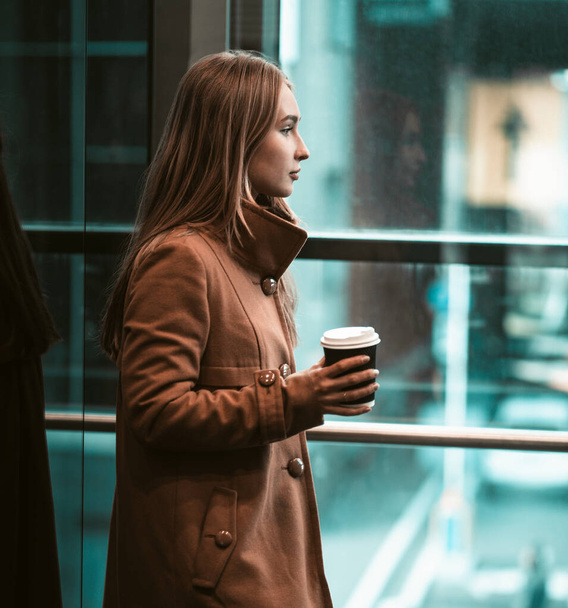 Young woman drinking coffee in business center. Long-haired European woman in warm coat holds paper cup looking out window - Foto, Imagem