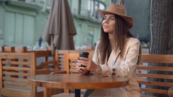 fiatal gyönyörű nő filckalapban ül egy asztalnál egy kávézóban az utcán. kávézás, eldobható papírpohár - Felvétel, videó