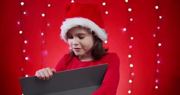 cute girl in a santa hat opens a gift on a red Christmas background - Imágenes, Vídeo