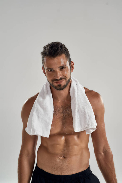 Vertical shot of a young handsome muscular man with towel on his shoulders resting after training, looking at camera and smiling while standing isolated over grey background - Фото, зображення