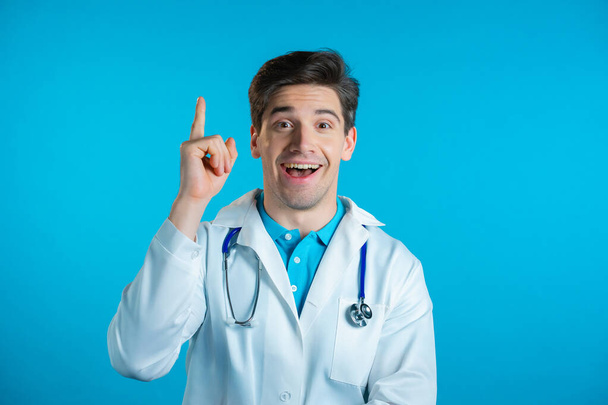 Portrait of doctor man in medical coat having idea moment pointing finger up on blue studio background. Smiling happy doc showing eureka gesture. - Fotó, kép