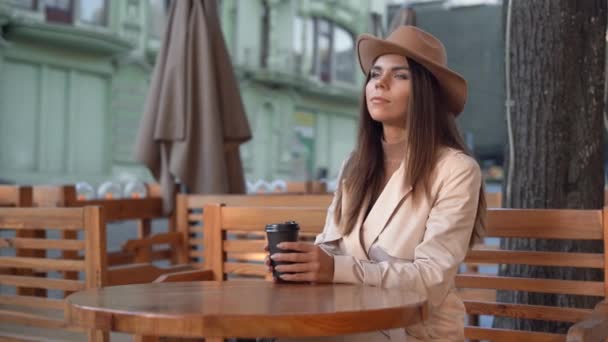 la bella donna giovane in un cappello sentito si siede a un tavolo in un caffè sulla strada. bere caffè, tazza di carta usa e getta - Filmati, video