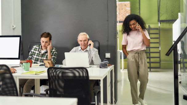 Aged man, senior intern talking on the phone while using laptop, sitting at desk, working together with other employees in modern office - Photo, image
