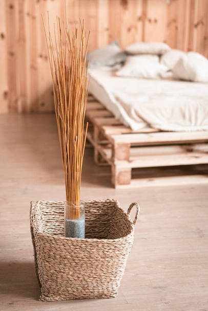 Natural home decor in wooden interior of bedroom. Bouquet of dried sticks in vase and wicker basket at floor - Фото, зображення