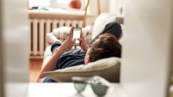 Teenage guy using his mobile phone while lying on the couch at home. The concept of smartphone addiction, apathy and frustration - Valokuva, kuva